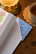 Open notebook with a blue fabric square on a wooden surface with a lit lamp and cup.